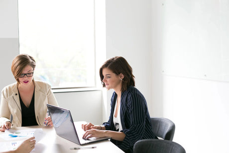 Two businesswomen in a modern office analyzing documents and working on a laptop during a meeting.